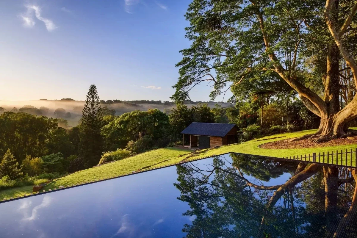 Reflection of trees and sky on a large, rectangular pool beside a grassy yard, a large tree, and a small wooden shed, overlooking a valley near Byron Bay.